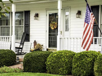 white colonial home decorated for fall with American flag flying from the front porch with rocking chairs